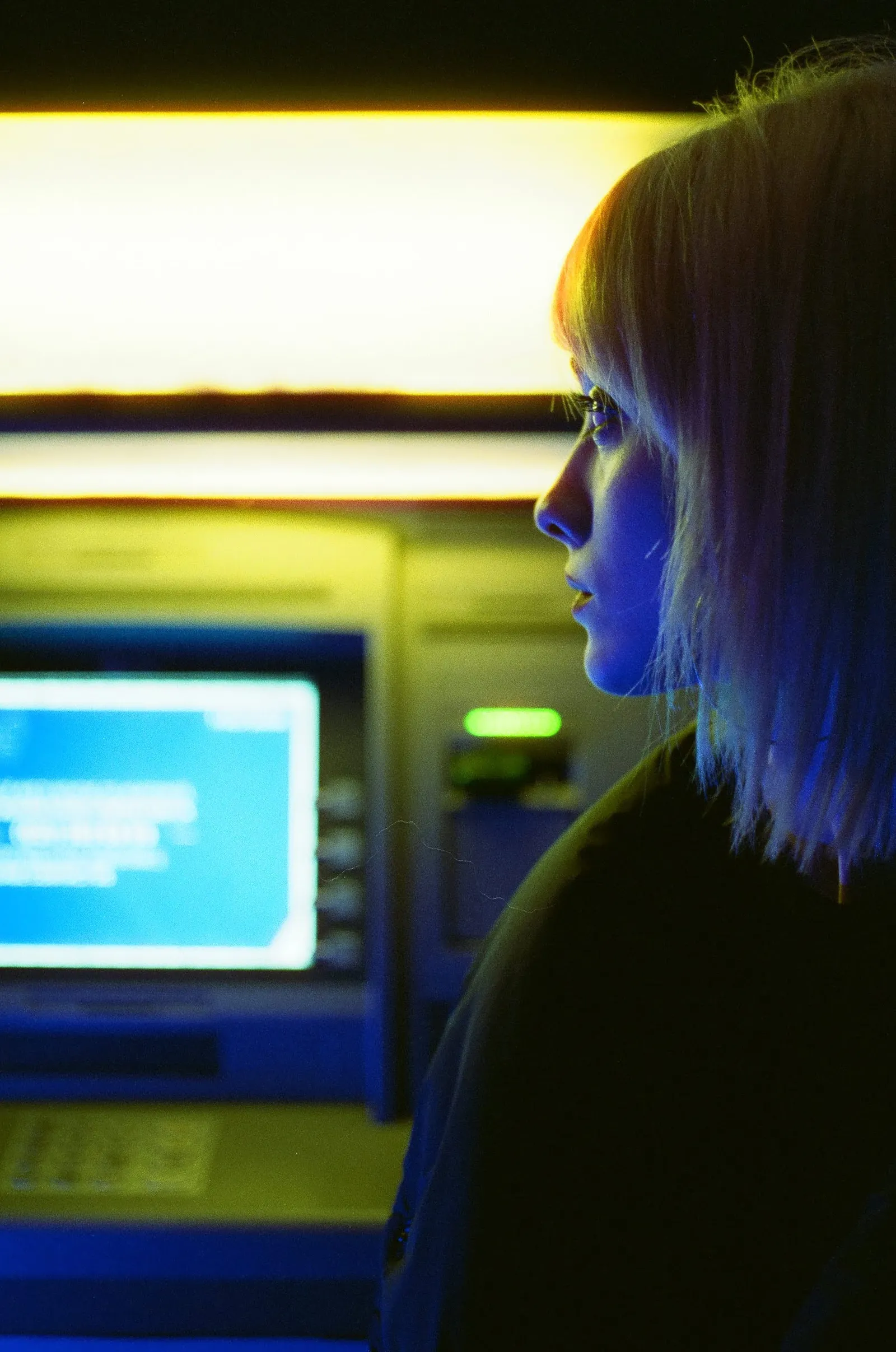 Oana in profile at a cash machine, blue screen light against warm tungsten