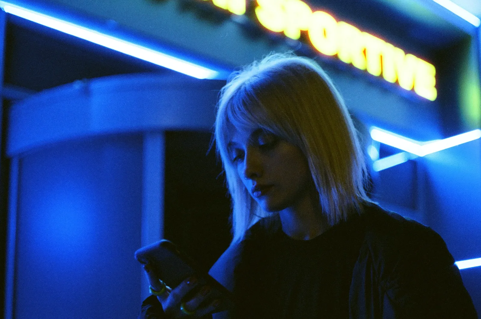 Oana lit blue by a 'Sportive' sign, looking down at her phone