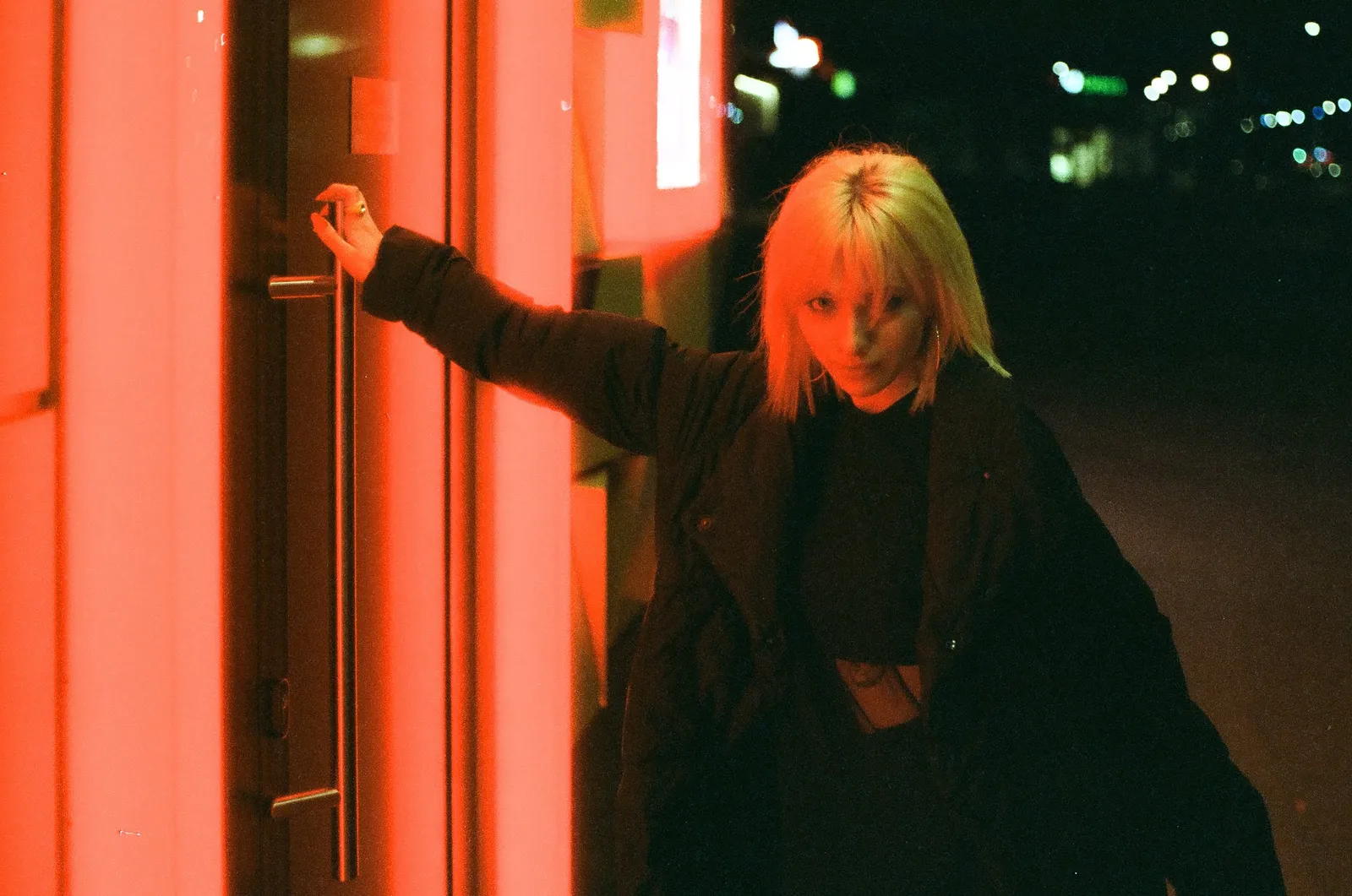 Oana leaning on a red-lit glass door, glancing back over her shoulder