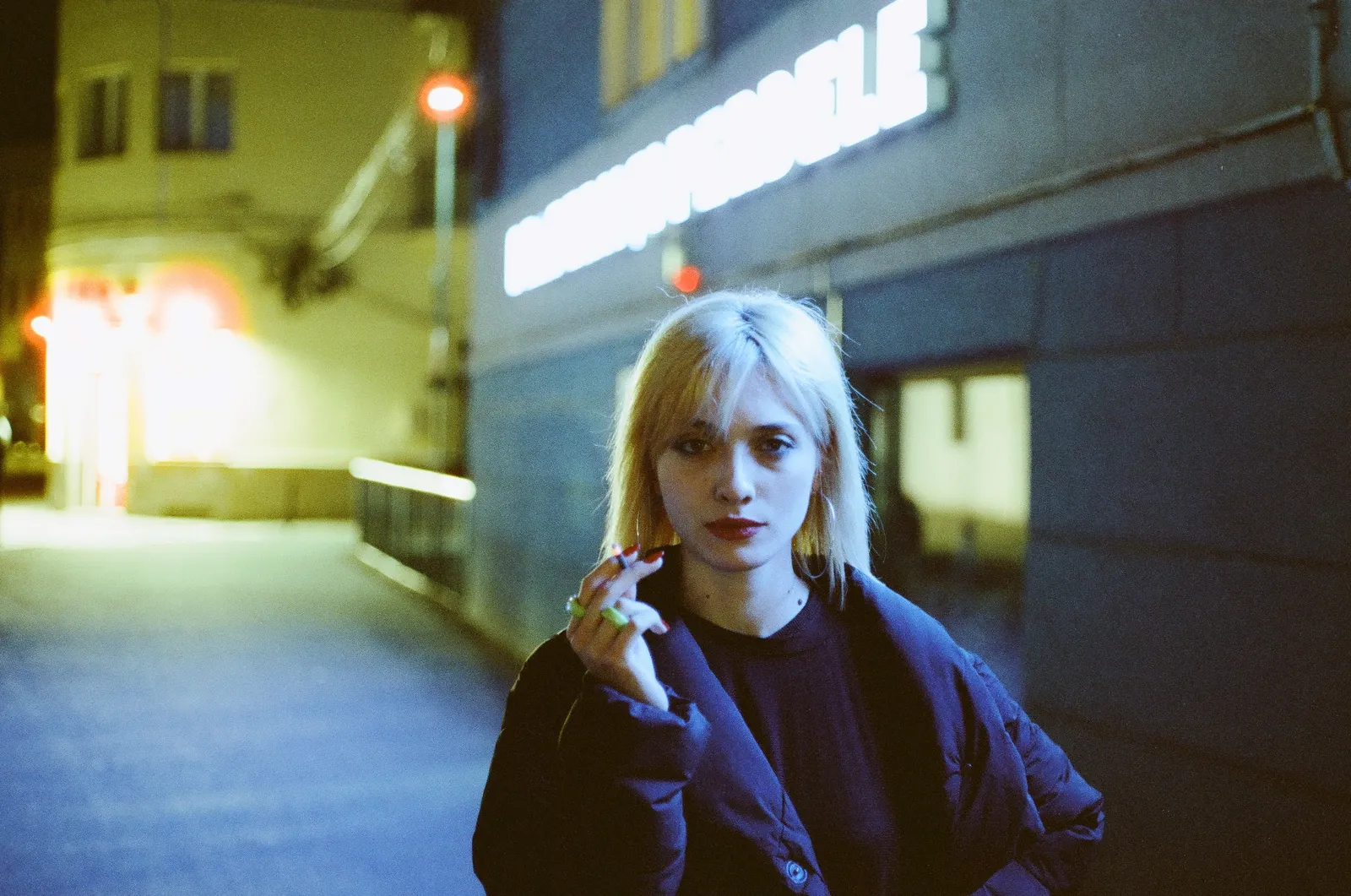 Oana looking straight into the lens under cool teal light, cigarette in hand