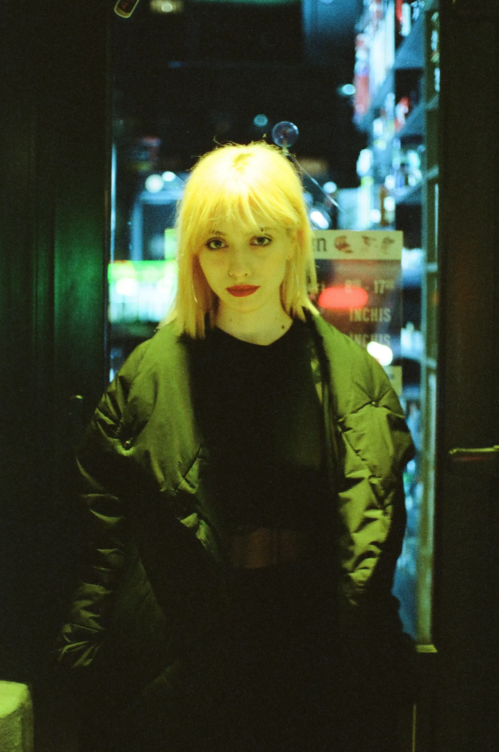 Oana in a doorway in an olive bomber jacket, a bar glowing behind the glass