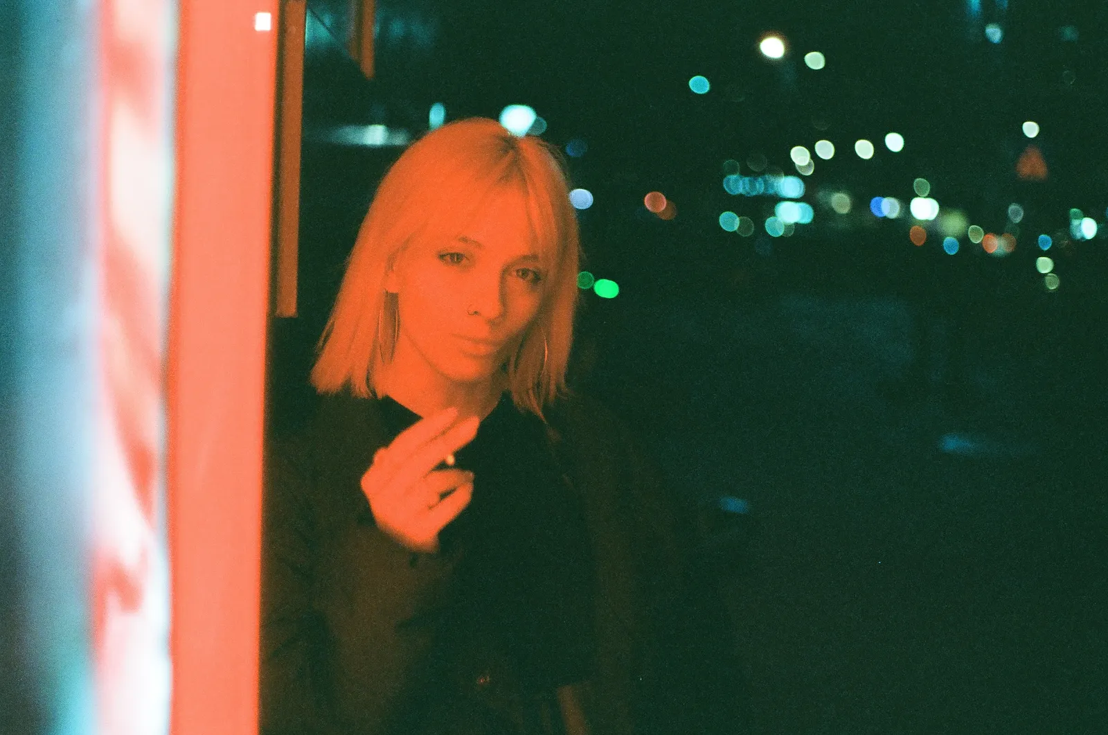 Oana lit by a red neon pillar, city lights melting into bokeh behind her