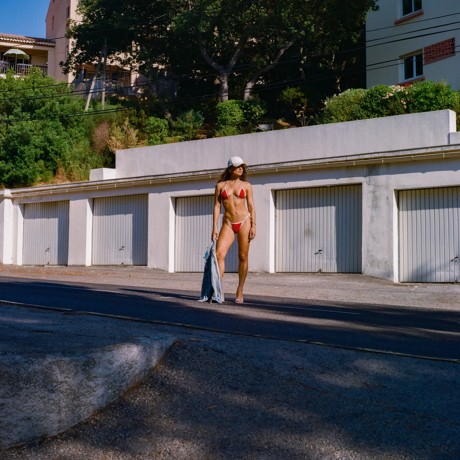 Model in a red bikini standing on an empty street in front of a row of white garages, denim jacket in hand