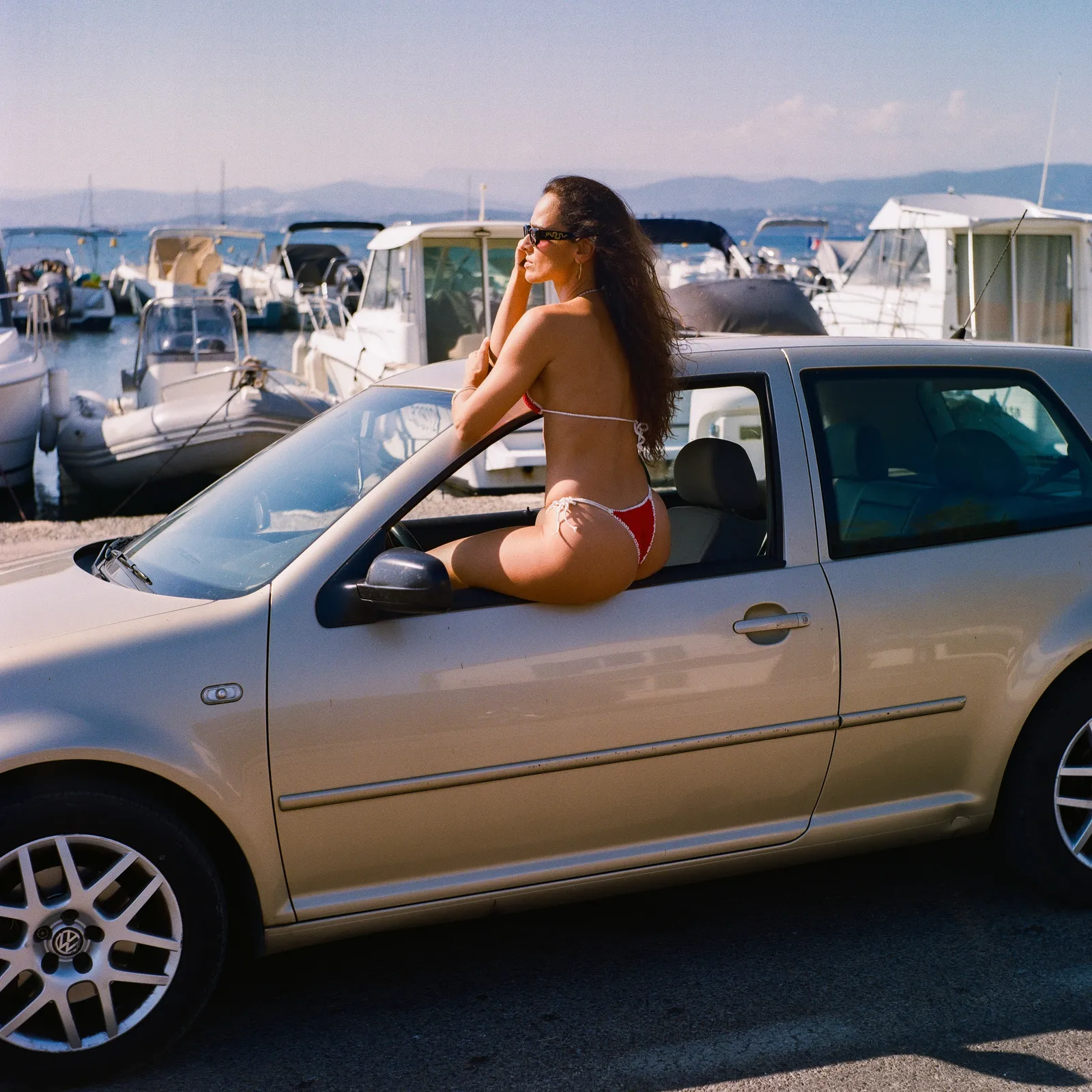 Model in sunglasses sitting in the open window of a champagne-coloured hatchback, boats in a harbour behind